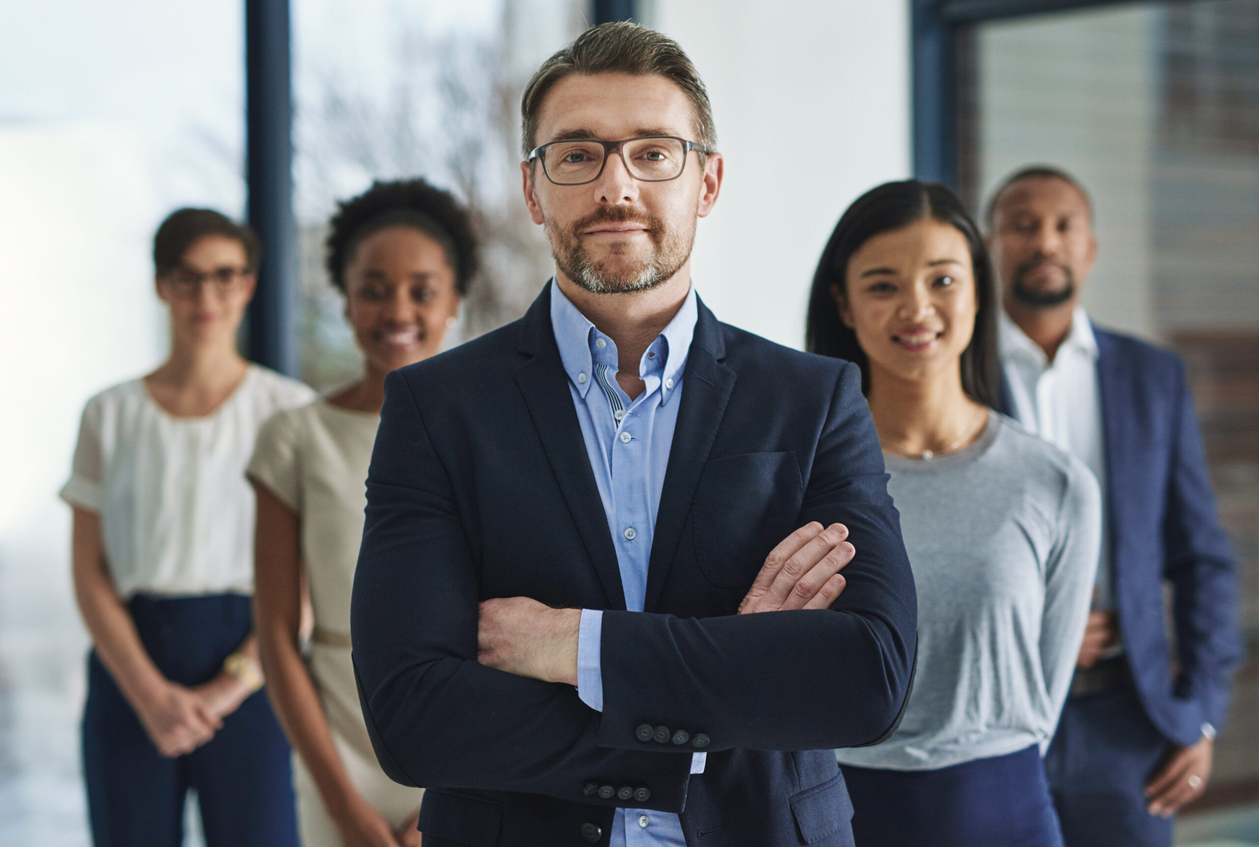 Homme d’affaires confiant en costume bleu, bras croisés, Homme d’affaires confiant en costume bleu, bras croisés, représentant le leadership et l’expertise en gestion comptable et financière, avec une équipe en arrière-plan
